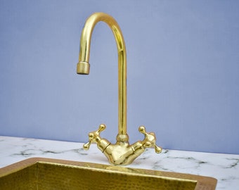 May include: A gold-toned kitchen sink faucet with two cross-shaped handles. The faucet is mounted on a white marble countertop and has a curved spout.