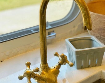May include: A gold-toned kitchen faucet with two cross-shaped handles. The faucet has a high arc spout and is mounted on a white countertop.