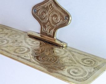 May include: A gold-colored metal shelf with an ornate design. The shelf is attached to the wall with a decorative bracket.