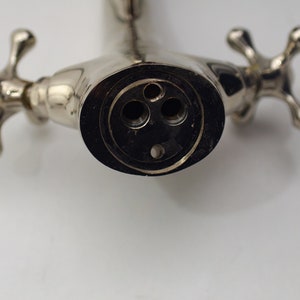 Pode incluir: Close-up de uma torneira cromada com duas al&ccedil;as em forma de cruz. A torneira tem tr&ecirc;s furos no bico, um grande e dois menores.