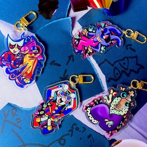 Può includere: Una collezione di portachiavi in acrilico colorati con chiusure color oro. I portachiavi presentano vari disegni di personaggi dei cartoni animati in colori vivaci come rosa, blu e giallo. Lo sfondo include carta blu strappata e coriandoli colorati.