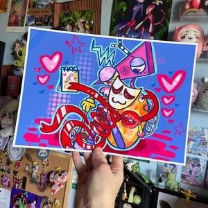 May include: A vibrant art print showcasing a cartoon character with a white face, red ribbons, and a pink triangle head. The background is blue with hearts and stars. The print is held by a hand, with various collectibles and art in the background.