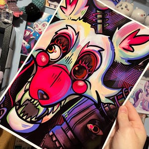 May include: A colorful art print featuring a stylized fox character with large eyes, a pink nose, and sharp teeth. The artwork uses a vibrant palette of pink, white, black, and blue, with intricate details and bold lines. The print is held by a hand.