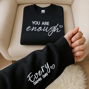 Peut inclure: Un sweat-shirt noir à manches longues avec le texte blanc "YOU ARE enough" sur la poitrine et une petite icône de cœur. La manche affiche le texte blanc "Every DAMN DAY" accompagné d'une autre petite icône de cœur.