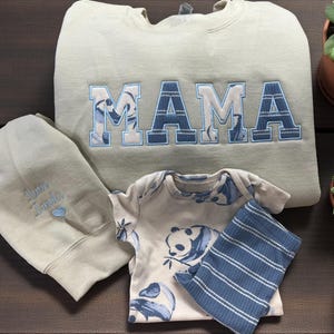 Op de afbeelding: Een beige sweater met ronde hals en het woord MAMA geborduurd in blauw-witte blokletters met patroon, een bijpassende beige romper met een blauwe panda-print, een blauw-wit gestreept geribbeld stoffen item en een beige muts.