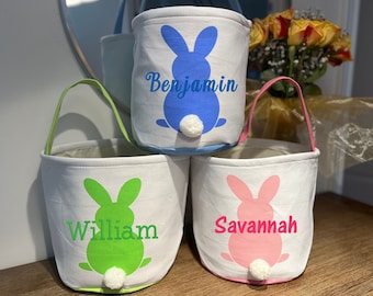 Puede incluir: Tres cestas de Pascua de lona blanca con siluetas de conejos y nombres personalizados. Las cestas son azul, verde y rosa con pompones blancos para las colas. Los nombres son "Benjamin", "William" y "Savannah".
