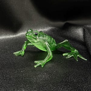 May include: A detailed, translucent green glass frog figurine. The frog is depicted with its legs extended, showing webbed feet. The figurine has a glossy finish and is set against a dark background.
