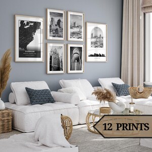 Puede incluir: Una sala de estar con un sofá seccional blanco y una mesa de café. La pared está decorada con impresiones en blanco y negro de rascacielos de la ciudad y mapas. Las impresiones están enmarcadas en marcos de madera natural. La habitación está decorada con una cesta de mimbre, un jarrón con hierba de la pampa seca y un portavelas de vidrio.