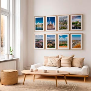 Puede incluir: Ocho fotografías de paisajes enmarcadas con marcos dorados se exhiben en una pared blanca. Las imágenes representan varios paisajes urbanos con cielos azules y cuerpos de agua. Un sofá blanco, una mesa de centro de madera y un otomán tejido están en primer plano.