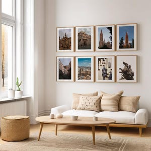 Puede incluir: Un interior de sala de estar con un sofá blanco, una mesa de café de madera y un puf tejido. La pared está decorada con ocho fotografías enmarcadas de varios paisajes urbanos y arquitectura.