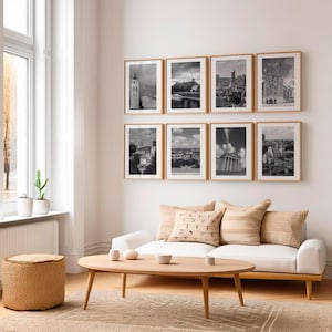 Puede incluir: Ocho fotografías enmarcadas en blanco y negro de paisajes urbanos cuelgan de una pared blanca sobre un sofá blanco con cojines beige. Una mesa de centro de madera y un otomán tejido están frente al sofá. Una ventana con una planta está a la izquierda.
