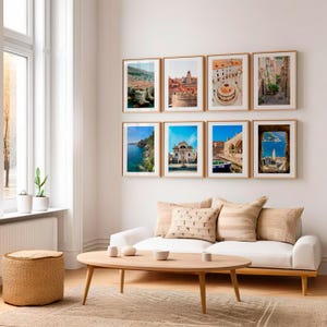 Puede incluir: Ocho fotografías de viajes enmarcadas con marcos dorados se exhiben en una pared blanca. Las imágenes representan escenas costeras y arquitectura. Un sofá blanco con cojines decorativos y una mesa de centro de madera están al frente.