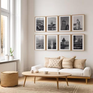 May include: A white couch with beige throw pillows sits in front of a white wall with eight black and white framed photographs of cityscapes and landscapes. A wooden coffee table sits in front of the couch.