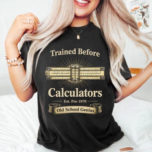 May include: Black t-shirt with the text "Trained Before Calculators" and "Old School Genius." The design features a slide rule with a sunburst effect. The shirt also includes the text "Est. Pre-1970."