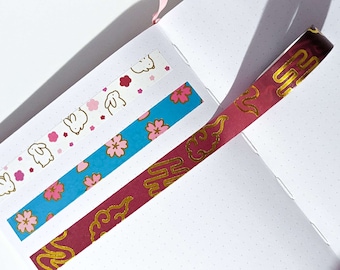 May include: Three strips of decorative washi tape applied to a white dotted notebook page. One strip features white rabbits and pink flowers, another shows pink cherry blossoms on a blue background, and the third is deep red with gold clouds.