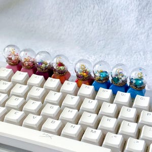 May include: A collection of custom keycaps featuring miniature snow globes. Each globe is filled with colorful decorations and sits atop a keycap base in various colors. The keyboard keys are white.