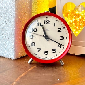 May include: A red alarm clock with a white face and black numbers sits on a wooden surface. The clock is next to a white gift box with a silver bow, a white lantern with a heart-shaped cutout, and a star-shaped candle.
