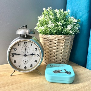 May include: A vintage silver alarm clock with a white face, next to a woven basket with greenery and a light blue Vespa box. The clock has black hands and numbers. The Vespa box has a scooter graphic.