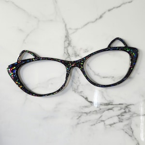 May include: Black cat-eye glasses with a glittery, multi-coloured speckled design. The frames have a unique cat ear shape at the top corners. The glasses are set against a white marble background.