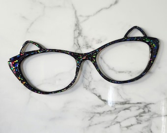May include: Black cat-eye glasses with a glittery, multi-colored speckled design. The frames have a unique cat ear shape at the top corners. The glasses are set against a white marble background.
