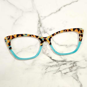 May include: Cat-eye glasses frames featuring a two-tone design. The upper portion of the rims displays a multi-colored leopard print pattern in shades of orange, black, and teal, while the lower portion consists of a solid, bright teal border.