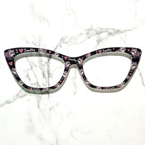 May include: Cat-eye eyeglass frames in black featuring a repeating pattern of small raccoons and the text I heart Jimothy with a red heart symbol. The frames have a distinct pointed outer edge and a smooth finish across the front surface.