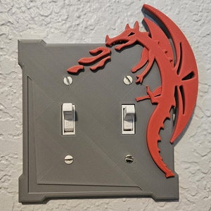 May include: A gray double light switch plate with a red dragon design. The dragon is facing right and has its wings spread out. The dragon is breathing fire.