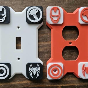 May include: Two decorative light switch and outlet covers. One is white with black accents and the other is orange with white accents. Each features superhero-themed emblems, including Iron Man, Thor's hammer, and Spider-Man.