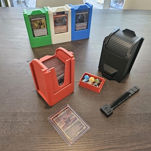 May include: A set of 3D printed card game accessories, including a red card holder, a black card shuffler, a small red dice tray with colorful dice, and a black card counter with the number "2" on it.