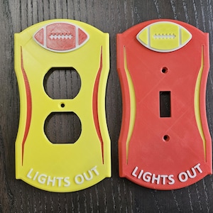 May include: Two light switch covers with a football design. The left cover is yellow with a red football and the right cover is red with a yellow football. Both covers have the text "Lights Out" printed on them.