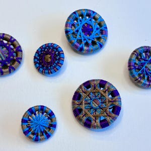 May include: A collection of seven handmade buttons in various sizes. The buttons feature intricate woven designs in shades of blue, purple, and gold. The buttons are arranged on a white surface, showcasing their detailed craftsmanship.