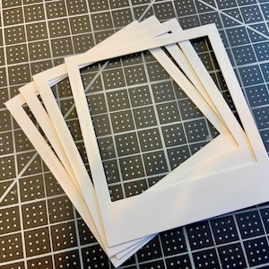 May include: A stack of white, square photo mats with a rectangular opening. The mats are made of thick paper and are stacked on a black cutting mat with white grid lines and angle markings.