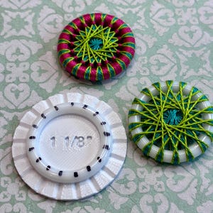 May include: Two circular buttons wrapped in vibrant magenta, lime green, and teal thread, alongside a white plastic tool with a fluted edge. The tool features the text 1 1/8 inches and black markings around the center to indicate size.