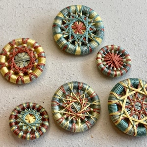 May include: A collection of six handmade, round buttons. Each button features a woven design with a central starburst pattern. The color palette includes shades of yellow, teal, and red, creating a visually appealing craft item.