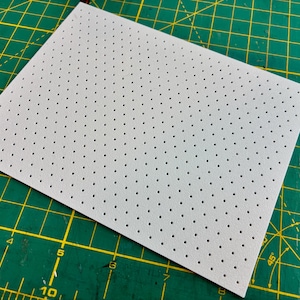 May include: A rectangular, light gray sheet of material with evenly spaced, small black perforations. The sheet is positioned diagonally on a green cutting mat with yellow grid lines, suggesting a crafting or design context.