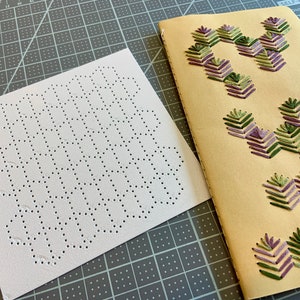 May include: A white fabric sheet with a grid of small holes, labeled "HEXIES", and a tan notebook with purple and green embroidery designs.