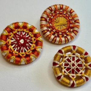 May include: Three round, embroidered buttons with a white, red, and yellow color scheme. The buttons have a circular design in the center and a concentric circle pattern around the center.