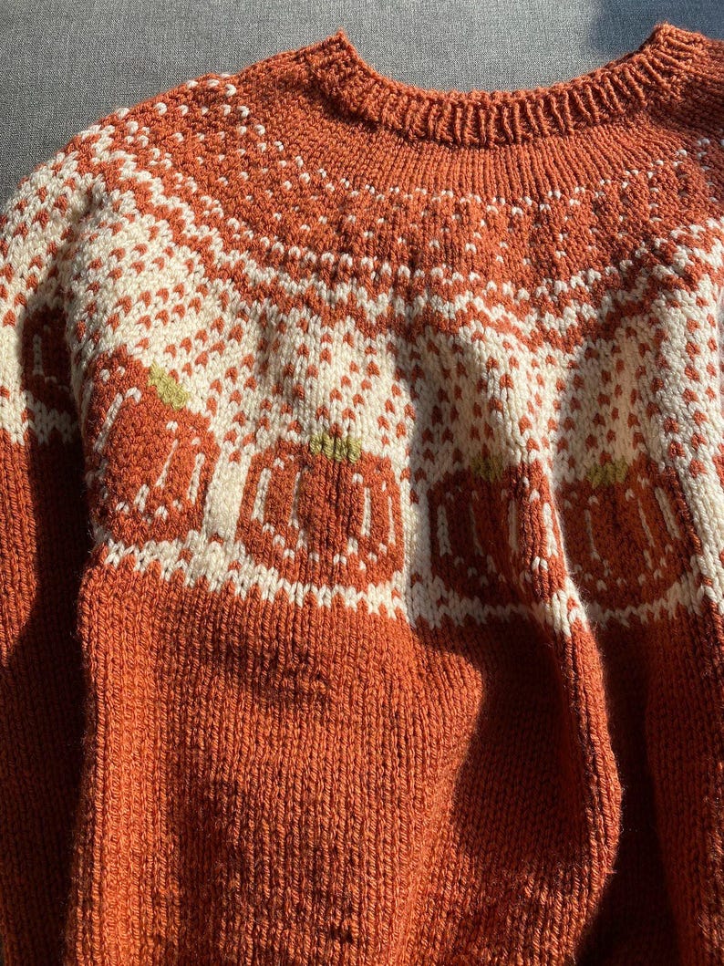 May include: A close-up of a brown knitted sweater with a white and orange pattern. The pattern features a repeating design of pumpkins.