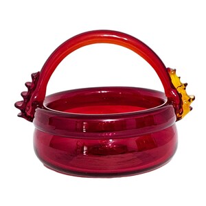 May include: A deep red glass basket featuring a smooth, rounded bowl and a high, arched handle. The handle is attached to the sides with decorative, amber-colored glass accents that resemble stylized fins or crests, adding texture to the piece.