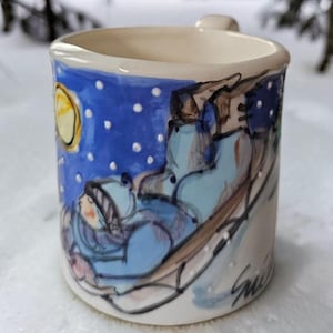 May include: A ceramic mug featuring a hand-painted scene of a person in a blue winter hat and coat riding a sled down a snowy hill under a yellow moon and blue night sky with white dots representing falling snow. The mug has a signature at the base.