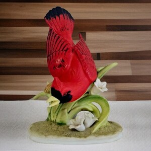 May include: A porcelain figurine of a red cardinal perched on a green leafy branch with white flowers. The cardinal is depicted with its head tilted down, as if it is looking at something.