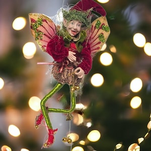 May include: A whimsical Christmas fairy ornament with a mischievous expression. The fairy wears a red velvet jacket, a green leafy headdress, and gold-trimmed wings. Its legs are green, and it has red shoes. The ornament is suspended by a string with small gold bells.