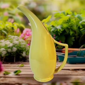 May include: A pale yellow ceramic pitcher with a long, curved spout and a handle. The pitcher has a smooth, glossy finish and a rounded base. The background is a blurred garden scene with green foliage and colorful flowers.
