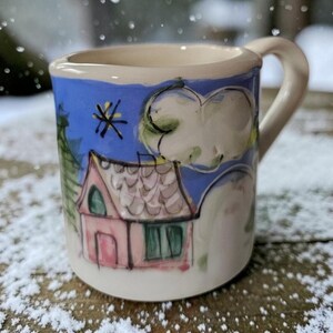 May include: A small ceramic mug with a cream-colored base featuring a hand-painted scene of a pink house with a green door and window, a green pine tree, and a yellow star against a blue sky, with white clouds and a curved handle on the side.