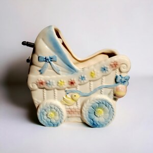 May include: A white ceramic baby carriage planter with blue and yellow floral details. The carriage has a blue ribbon and a yellow bird on the side.