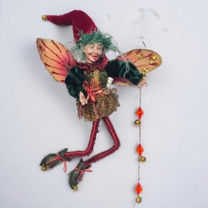 May include: A whimsical fairy figurine with a red velvet hat and green hair. The fairy wears a green and gold outfit with red legs and wings. It holds a string of orange beads and gold bells, perfect for holiday decor.