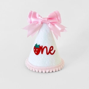 May include: White felt party hat with a pink satin bow, a red strawberry, and the word "one" in red. The hat has a pink pom-pom trim.
