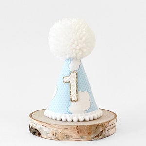 May include: A blue and white party hat with a white pom-pom on top. The hat has a white cloud pattern and a gold glitter number "1" on the front.