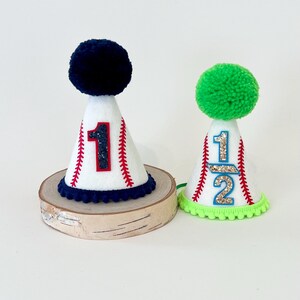 May include: Two white felt baseball-themed party hats with red stitching and a number on the front. The hat on the left has a navy blue pom-pom and the number "1". The hat on the right has a green pom-pom and the number "1/2".