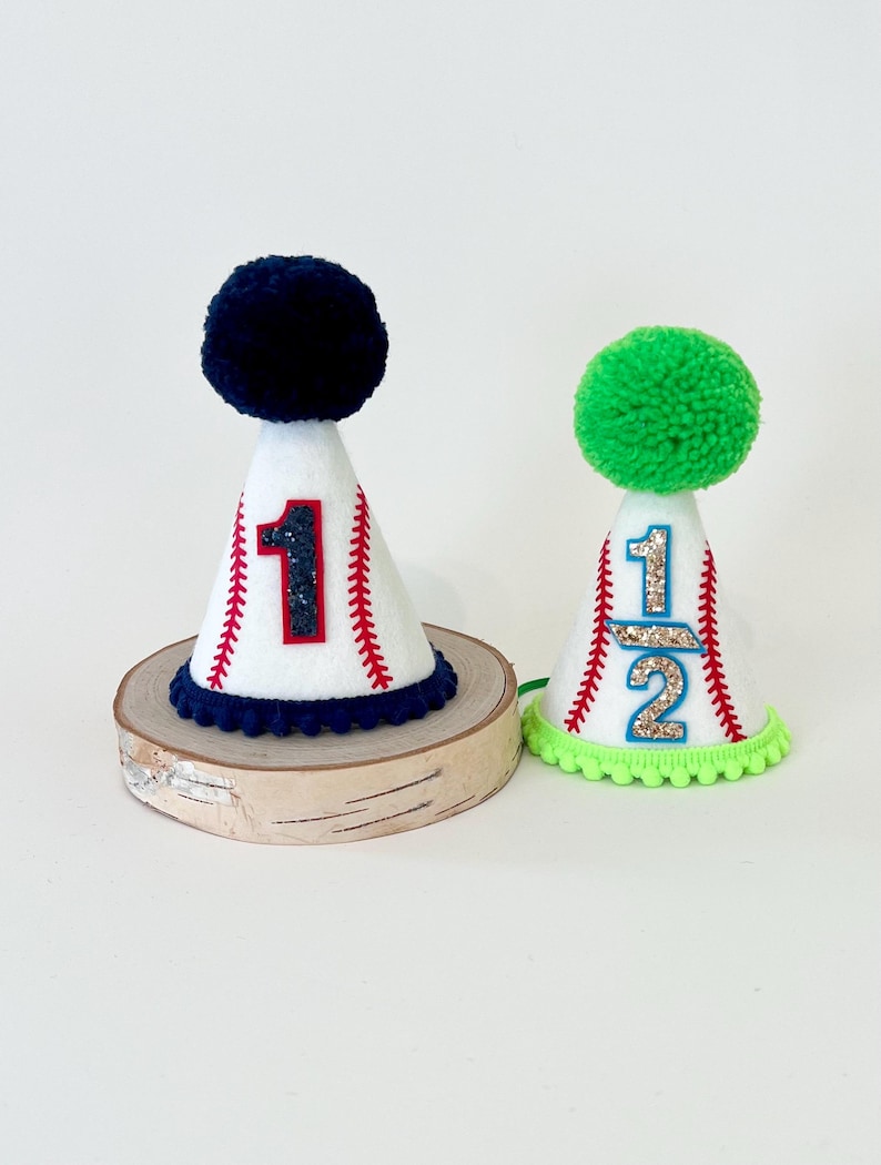 May include: Two white felt baseball-themed party hats with red stitching and a number on the front. The hat on the left has a navy blue pom-pom and the number "1". The hat on the right has a green pom-pom and the number "1/2".